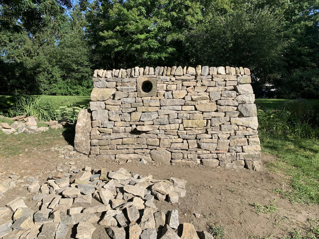 A newly built dry stone wall on a lawn, with a carved stone block showing a round hole set into it as a feature, loose stone piled in the foreground