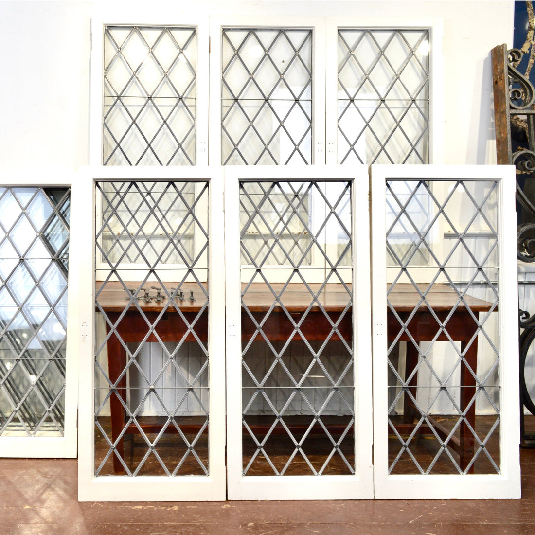 Tall white-framed leaded windows with a diamond lattice, standing in two rows against a white wall, a wooden work table visible through the glass