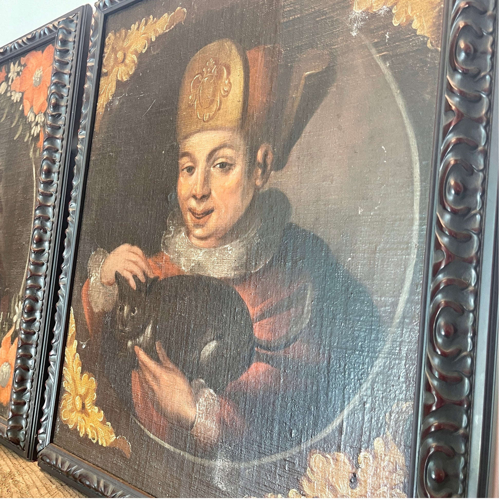 A close-up of a framed antique-style portrait painting showing a man in a gold hat and red robe holding a black cat, with ornate dark wood framing and partial adjacent panels visible.