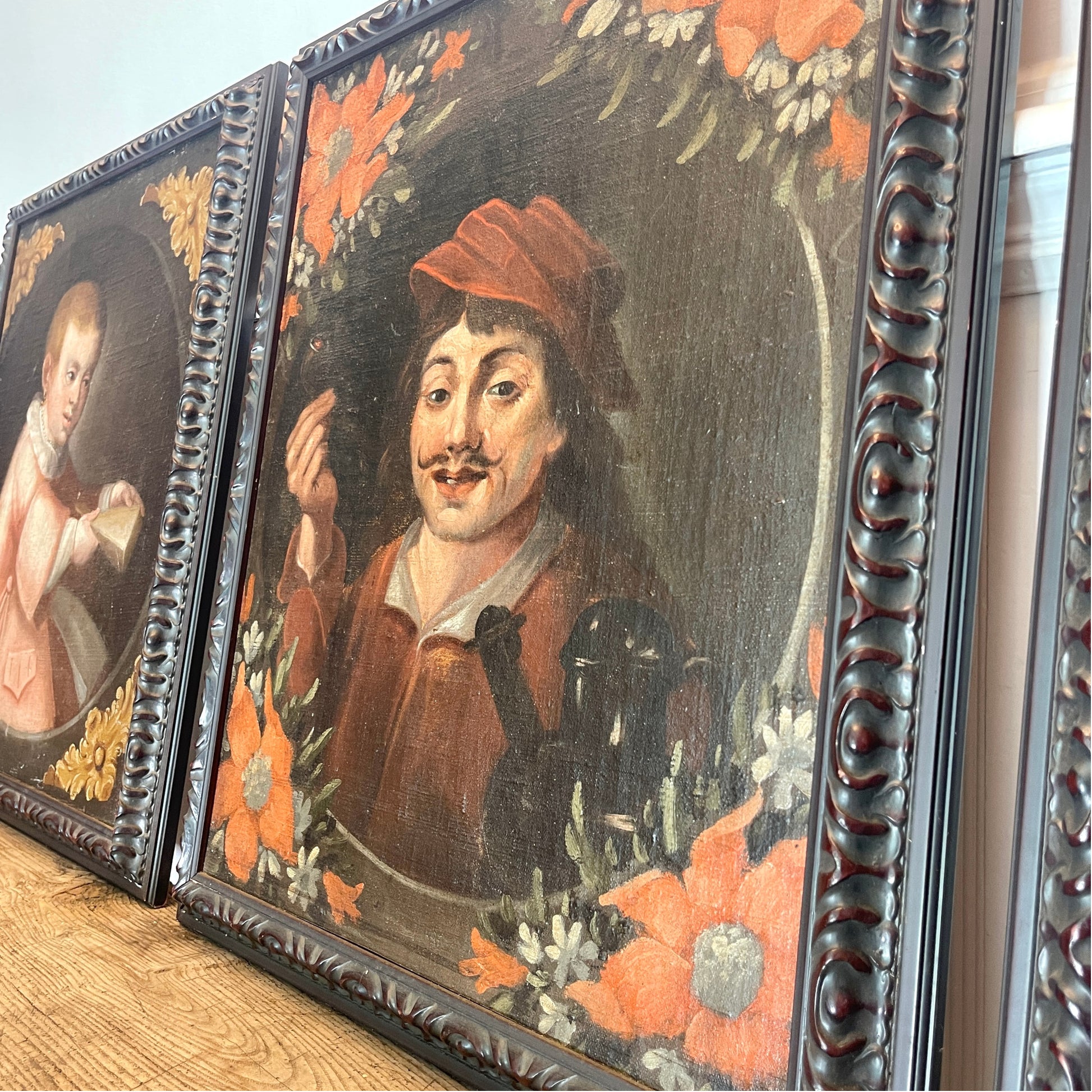A close-up of a framed vintage oil painting depicting a man in a red hat and brown clothing holding a pipe, urrounded by orange flowers, with another framed portrait partially visible on the left.