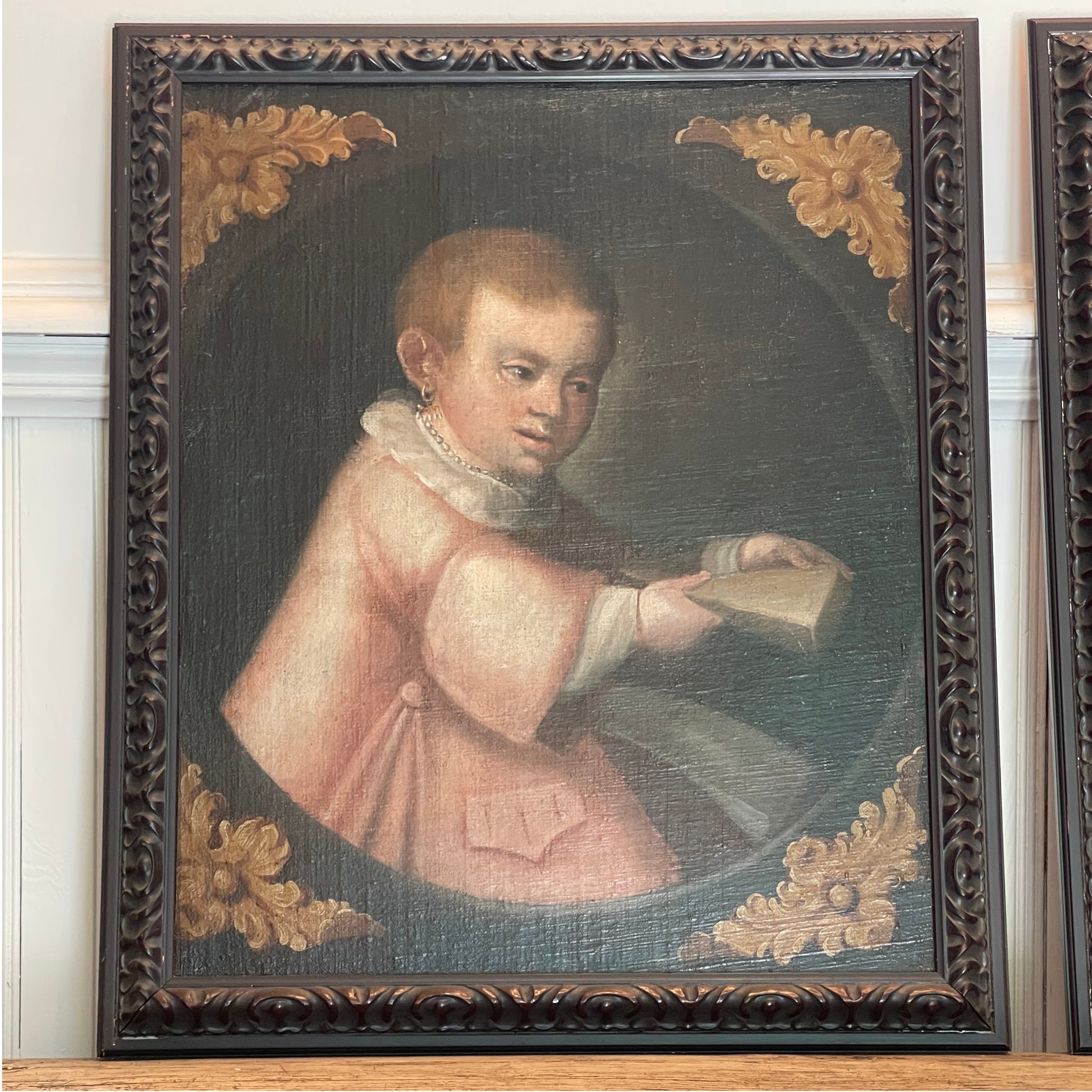 A framed antique-style portrait painting of a young boy in a pale pink robe holding a book, set in an ornate dark brown carved frame with gold corner details, displayed against a white paneled wall.