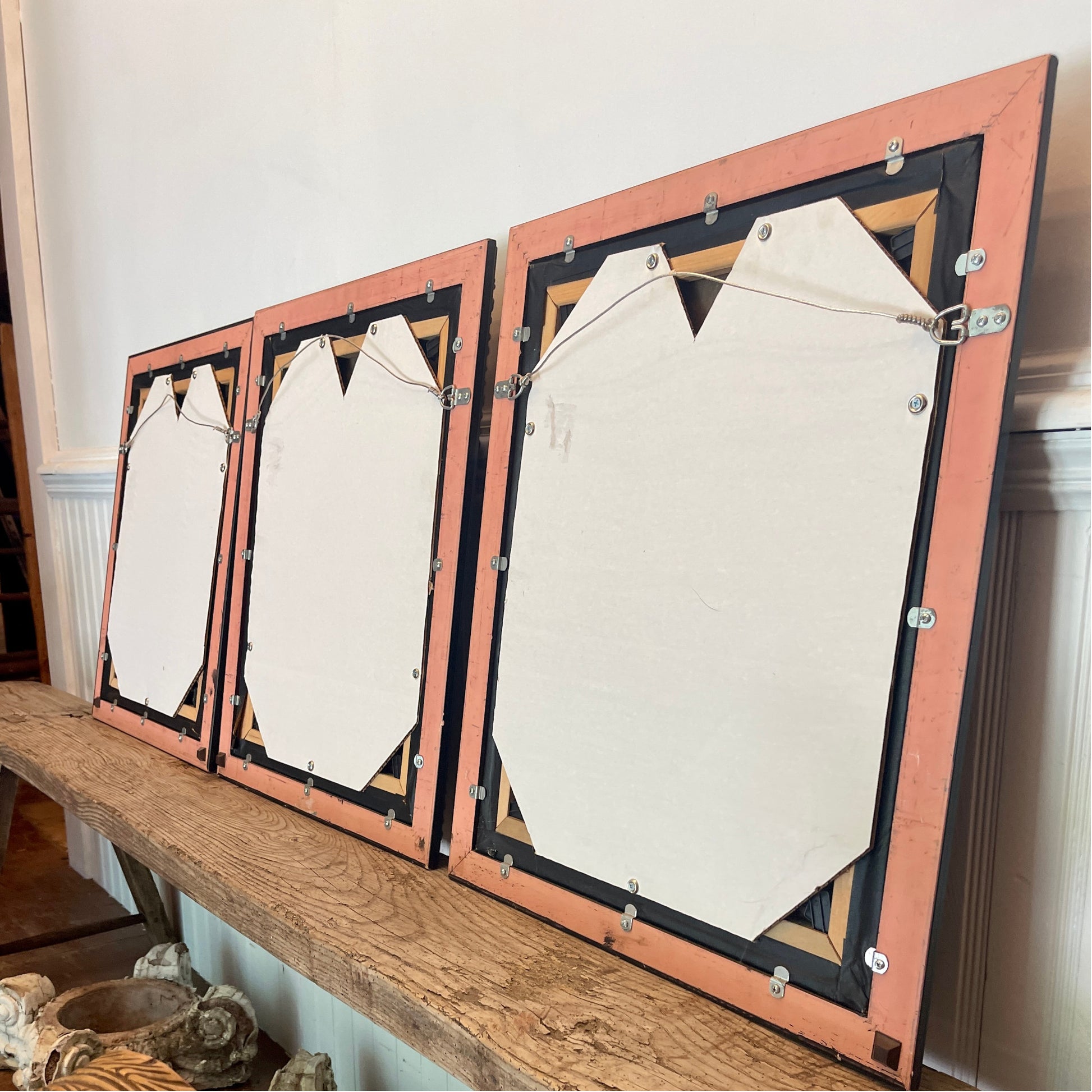 Three framed canvases displayed side-by-side on a rustic wooden shelf in a home interior. The canvases are shown from the back showing the level of restoration.