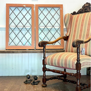 A vintage upholstered wooden chair with striped fabric in beige, pink, and green, positioned next to two tall wooden window frames with diamond-patterned glass on a white paneled wall.
