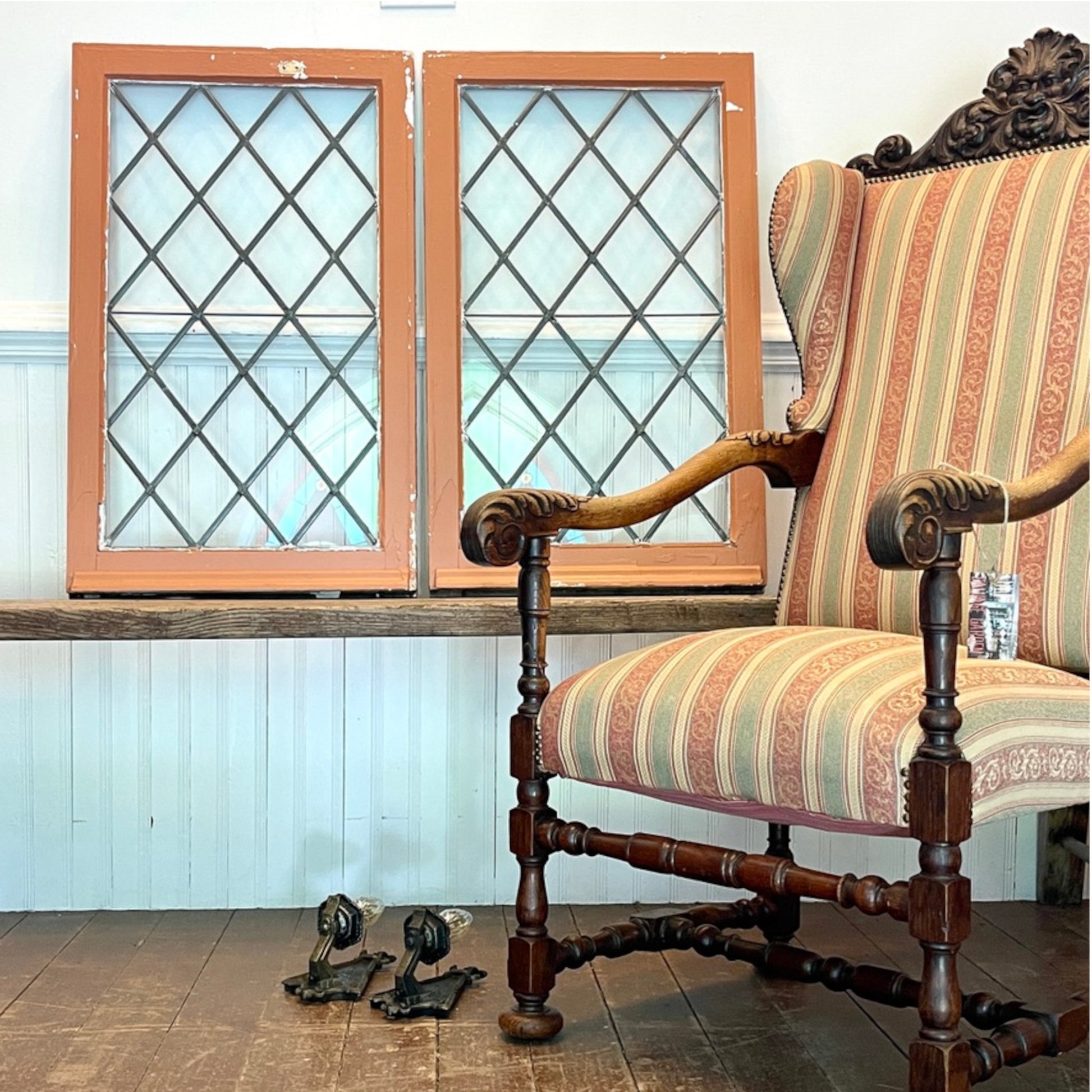 A vintage upholstered wooden chair with striped fabric in beige, pink, and green, positioned next to two tall wooden window frames with diamond-patterned glass on a white paneled wall.