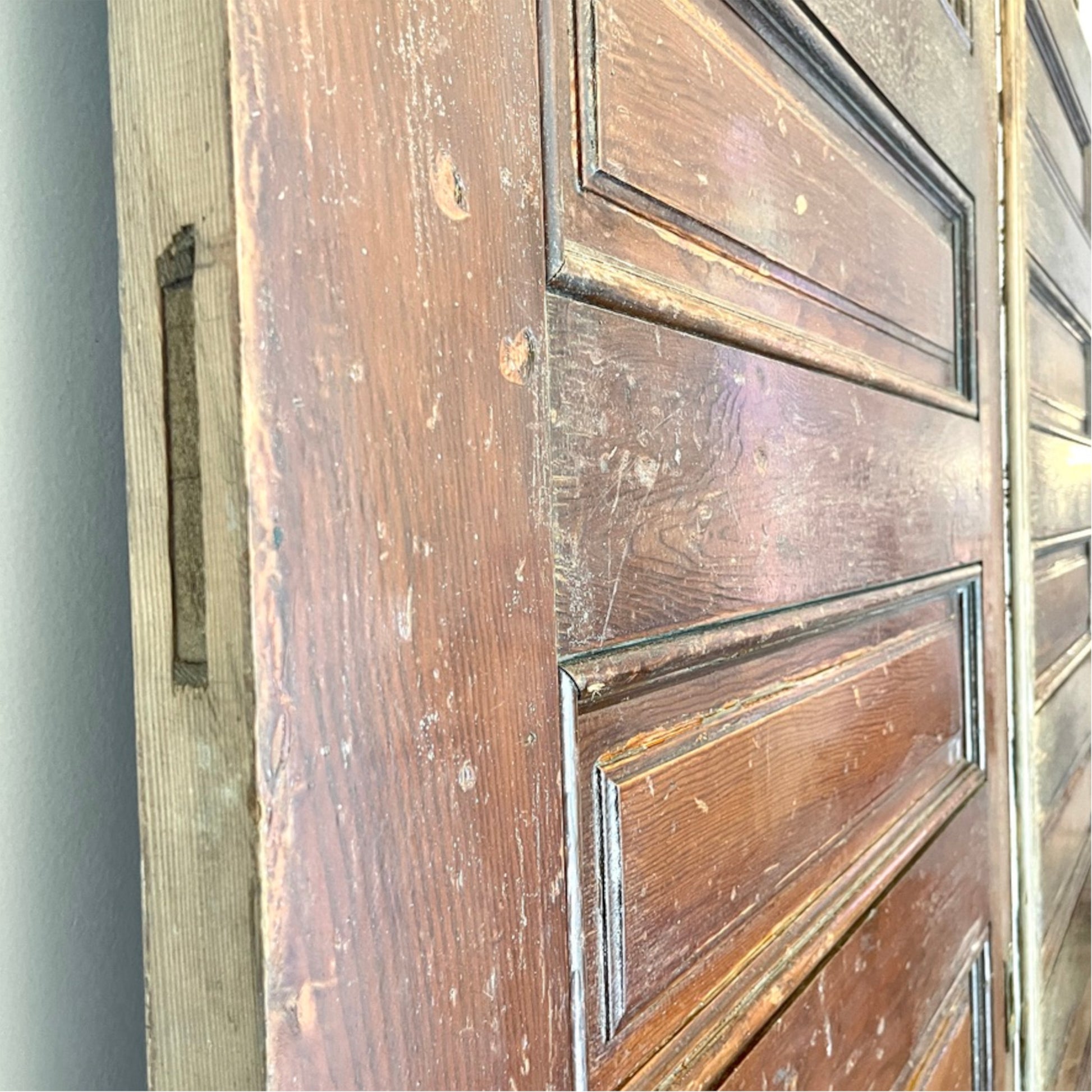 A close-up of a distressed medium-brown wooden door with raised panel molding and visible wear, scratches, and peeling paint, set against a neutral gray wall.