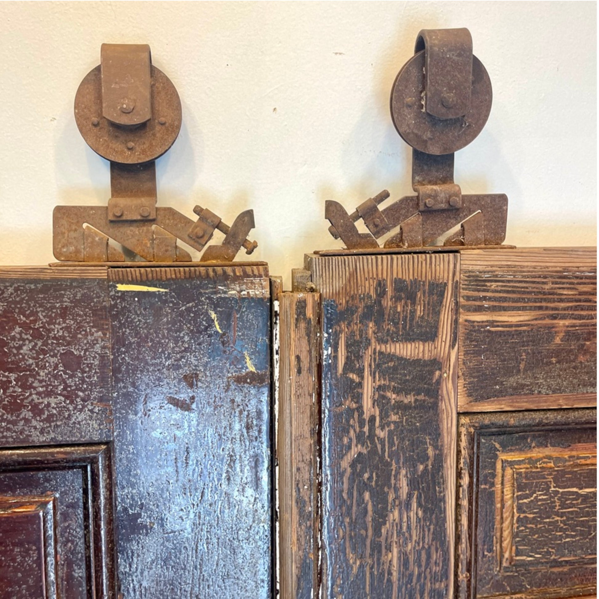 A pair of vintage dark brown wooden doors with heavy rusted metal roller hardware mounted above the top edges, set against a plain off-white wall.