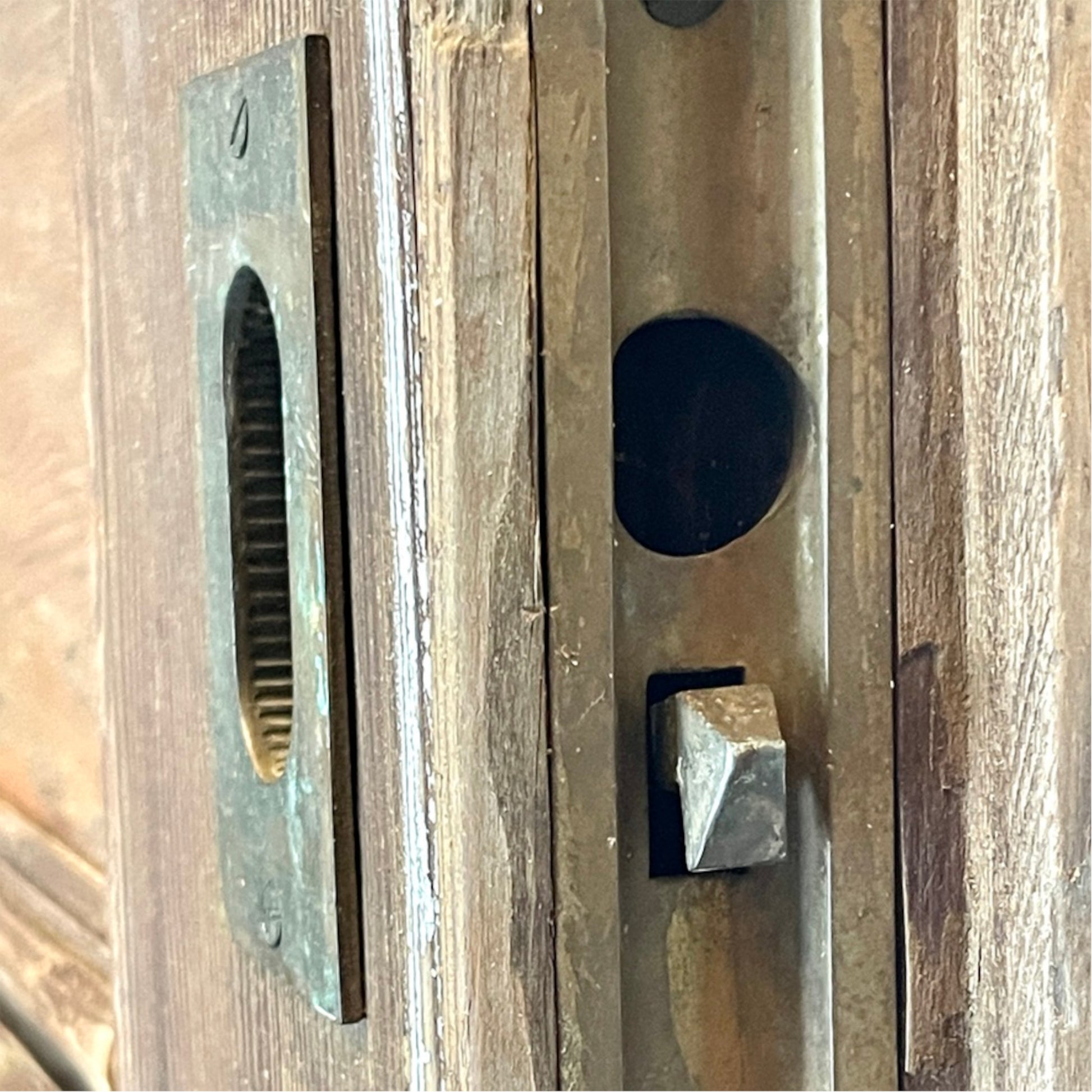 Close-up of a antique metal door lock mechanism with a rectangular slot plate and a square thumb turn on a weathered wooden door.
