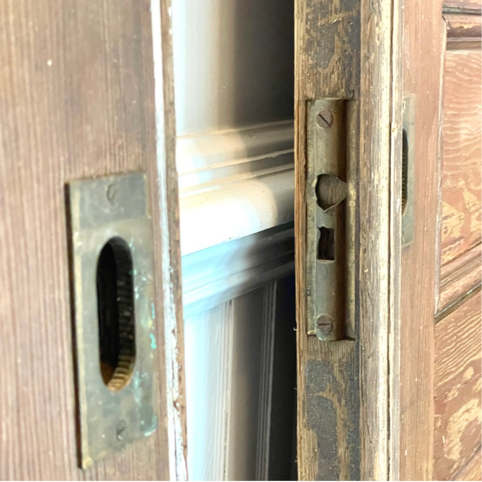 Close-up of a weathered wooden door with a brass or bronze oval door lock and a matching deadbolt on the right side, showing the latch mechanism and a glimpse of the interior.