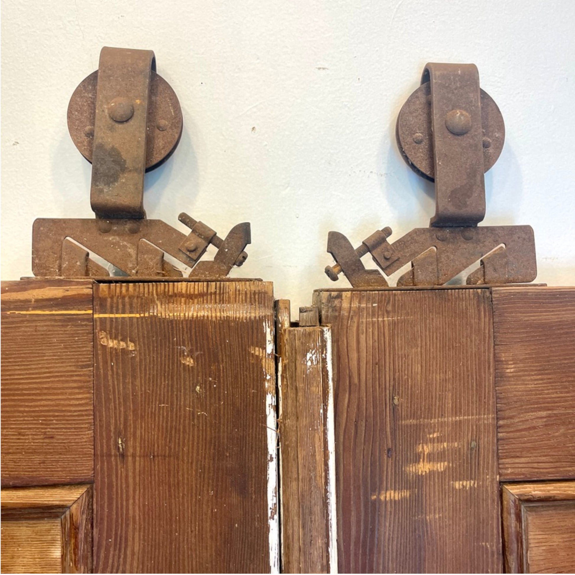 A pair of rustic, rusted metal pulley door hardware assemblies mounted on the top corners of weathered brown wooden barn doors against a light off-white wall.