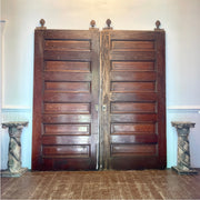 A pair of tall, dark brown wooden double doors with raised rectangular panels and small finials at the top, standing in a room with a wood floor and pale walls.