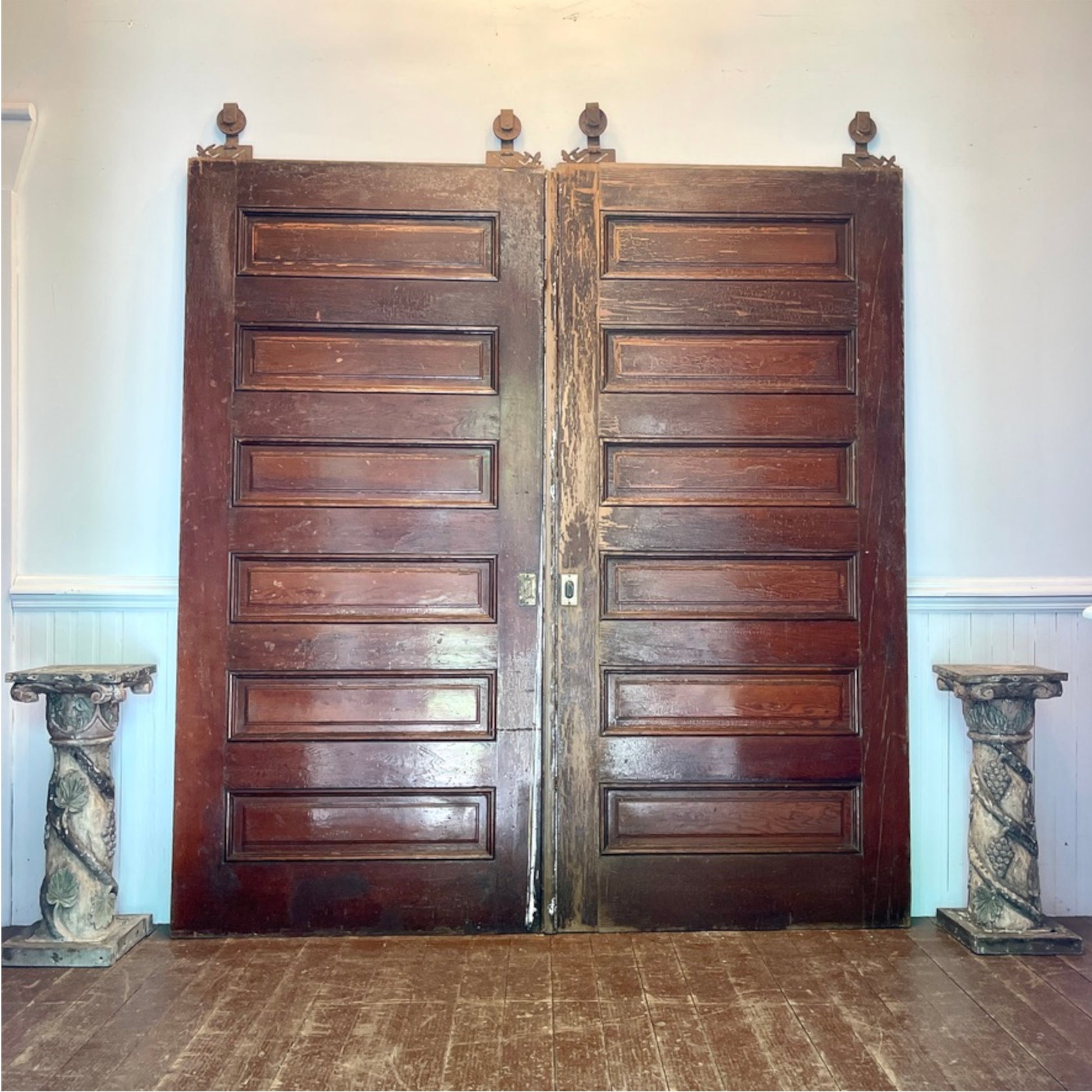 A pair of tall, dark brown wooden double doors with raised rectangular panels and small finials at the top, standing in a room with a wood floor and pale walls.