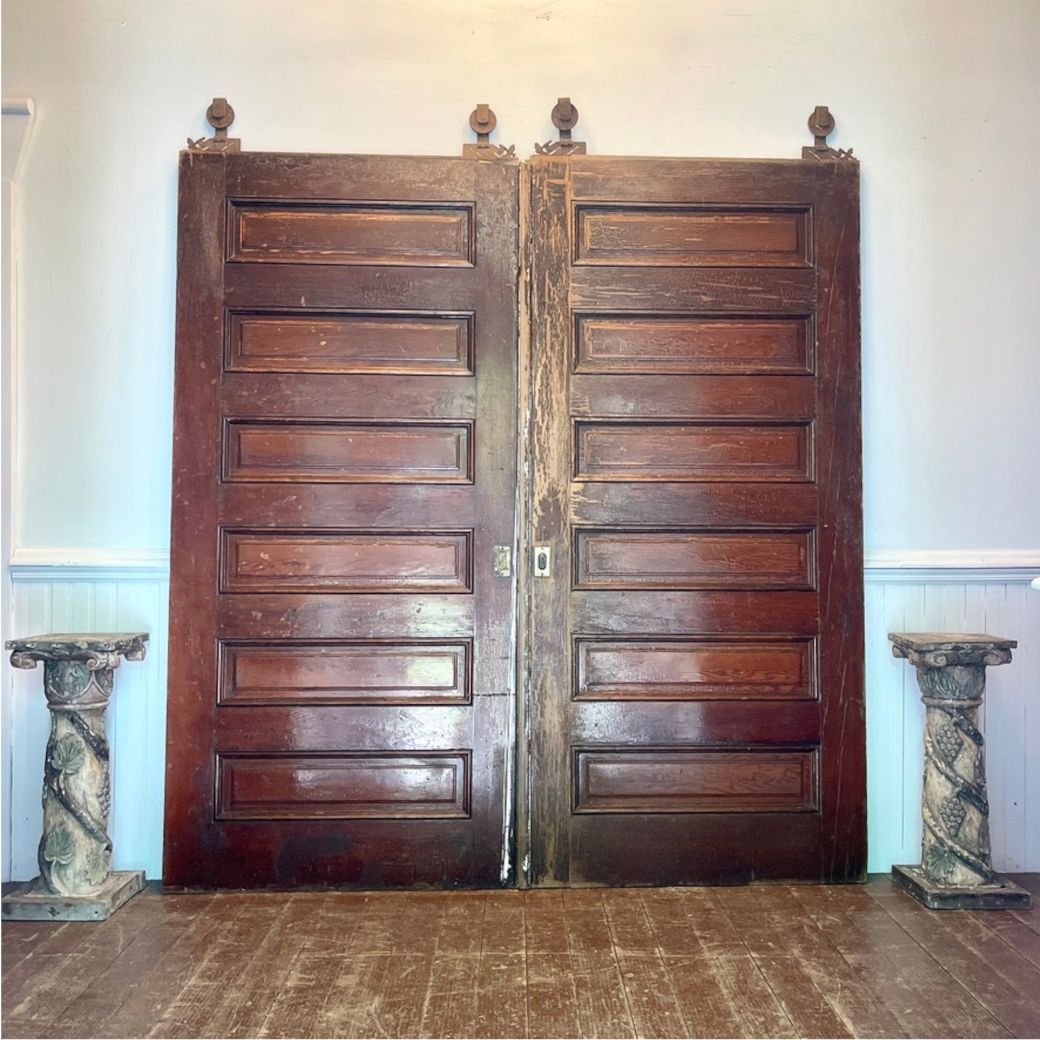 A pair of tall, dark brown wooden double doors with raised rectangular panels and small finials at the top, standing in a room with a wood floor and pale walls.