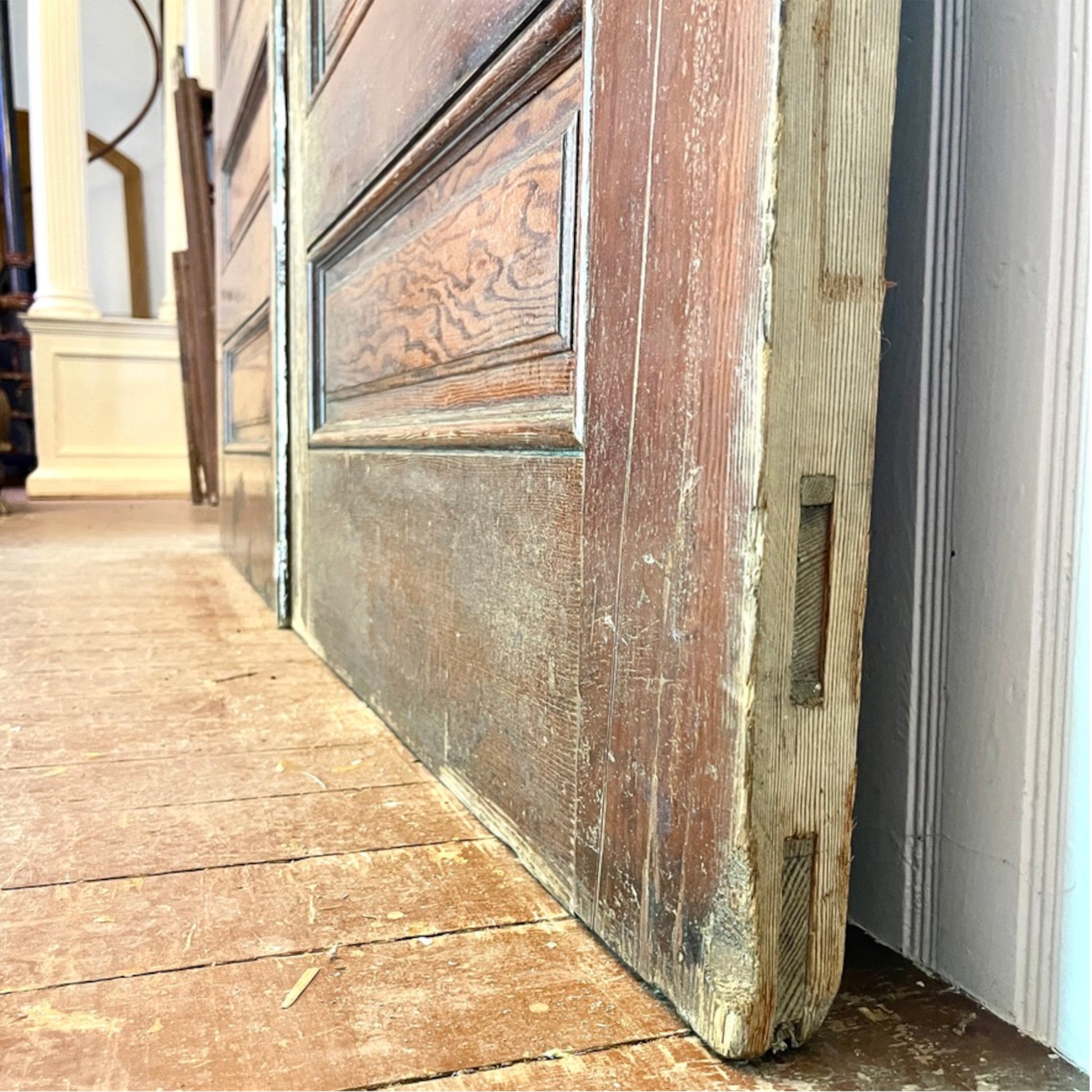 An antique wooden door panel with a distressed finish, leaning against a white wall on a wooden floor.