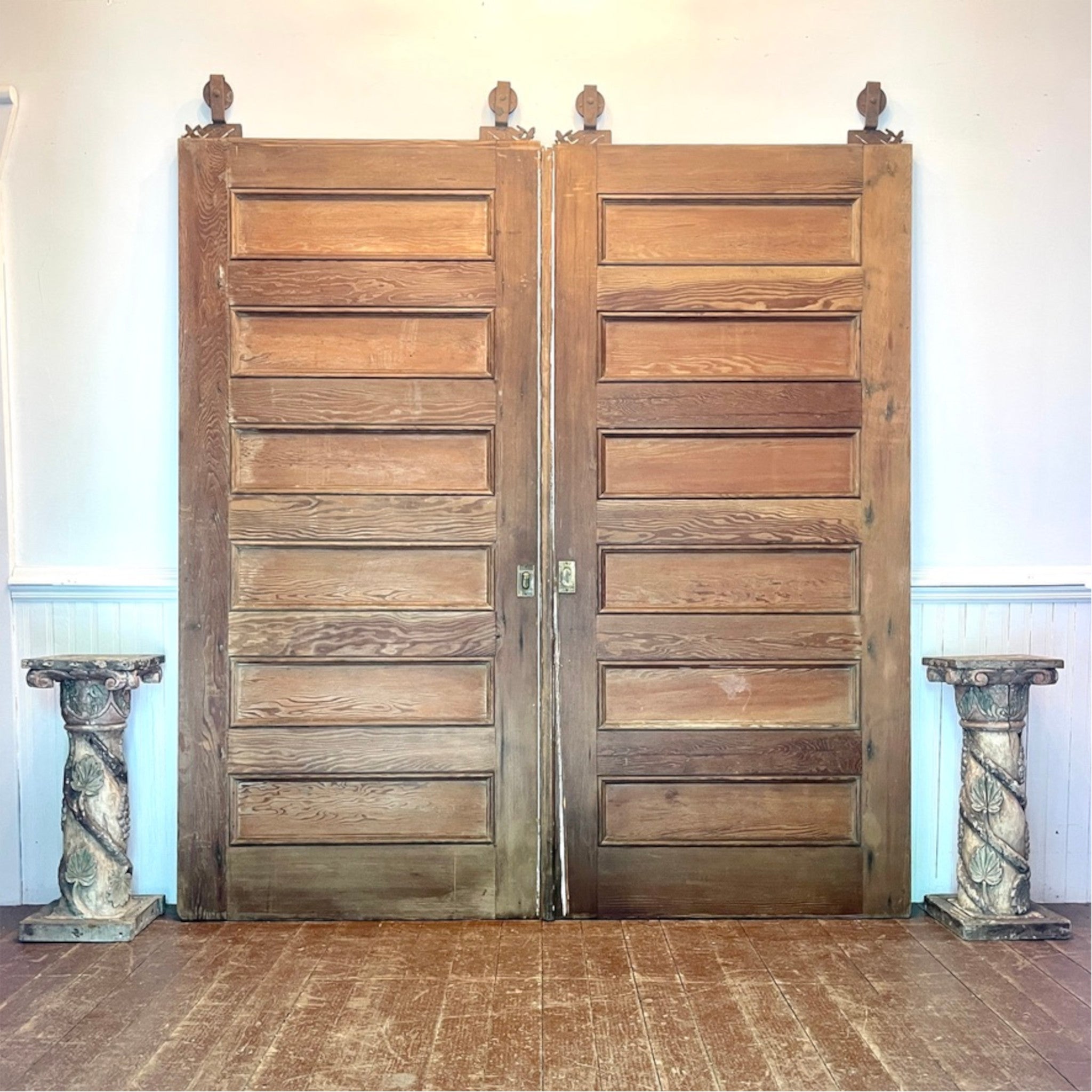 A pair of tall, rustic double wooden doors with a paneled design and original gliders at the top, standing in a room with a wood floor and white wainscoting.