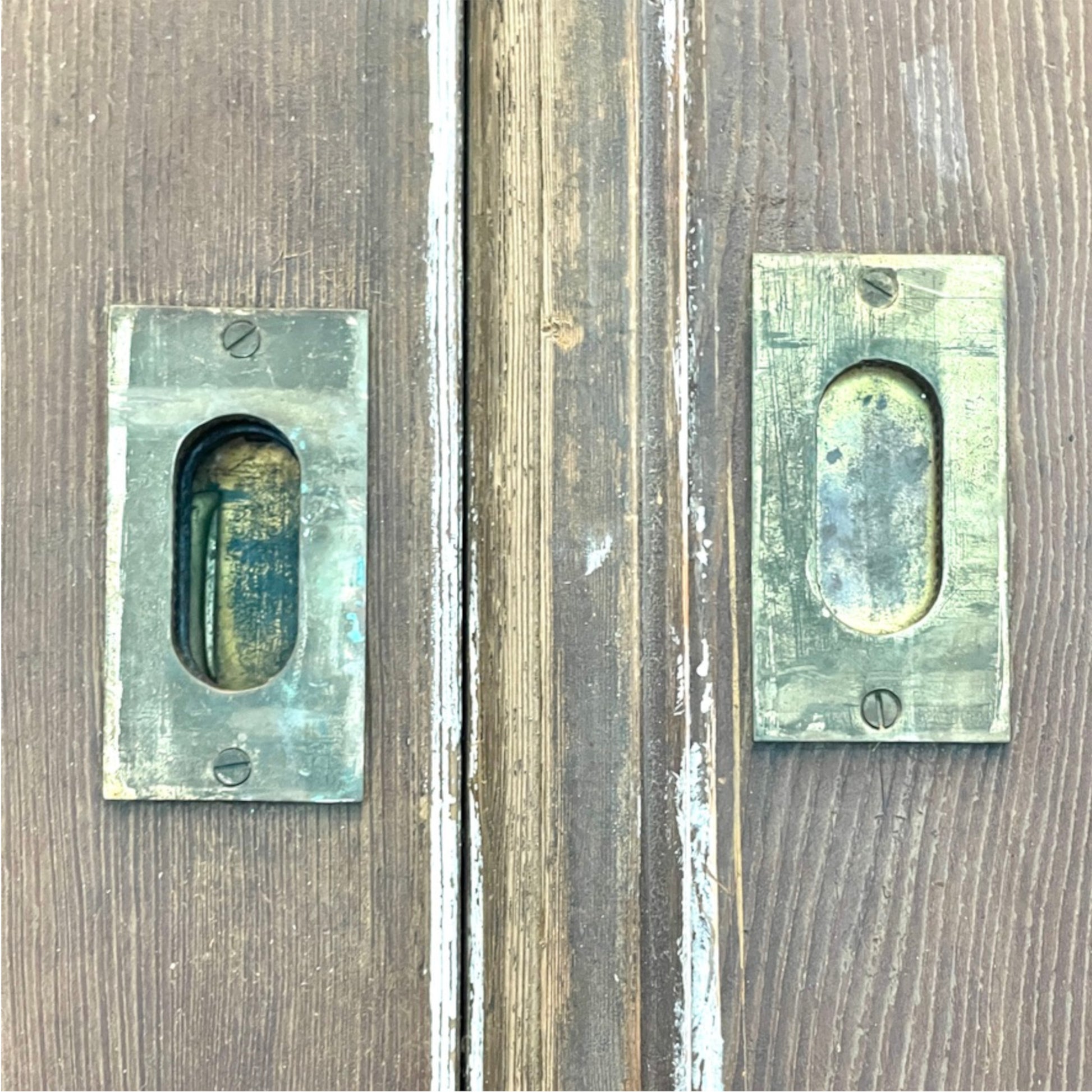Two vintage rectangular metal door pull handles with oval cutouts, mounted on weathered brown wooden doors.